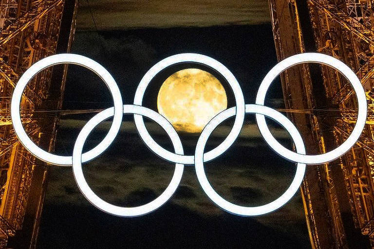 A imagem mostra os aros olímpicos iluminados pendurados na Torre Eiffel à noite. Ao fundo, a lua cheia aparece centralizada entre os aros, criando um efeito visual impressionante. A estrutura metálica da torre é visível, destacando-se contra o céu noturno.