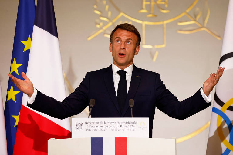 Um homem de terno escuro e gravata está discursando em um pódio com os braços abertos. Atrás dele há duas bandeiras, uma da França e outra da União Europeia. O pódio tem um emblema e uma placa com texto em francês, além de uma faixa com as cores da bandeira francesa.