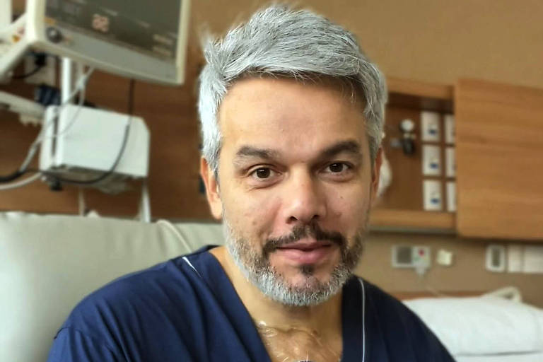 A imagem mostra um homem de cabelos grisalhos e barba sentado em um quarto de hospital
