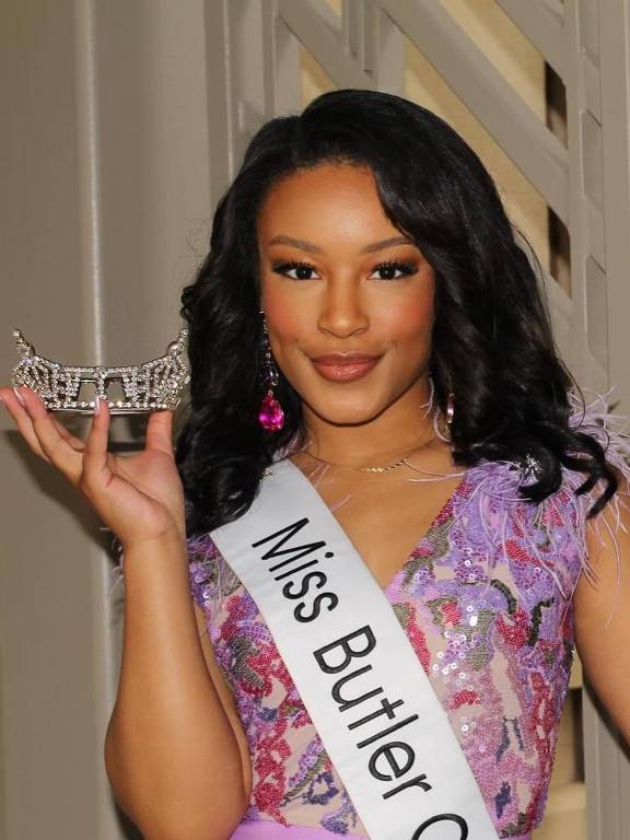 Uma mulher está posando para uma foto, segurando uma coroa de concurso de beleza em uma das mãos. Ela está usando uma faixa que diz 'Miss Butler County' e um vestido lilás com detalhes brilhante