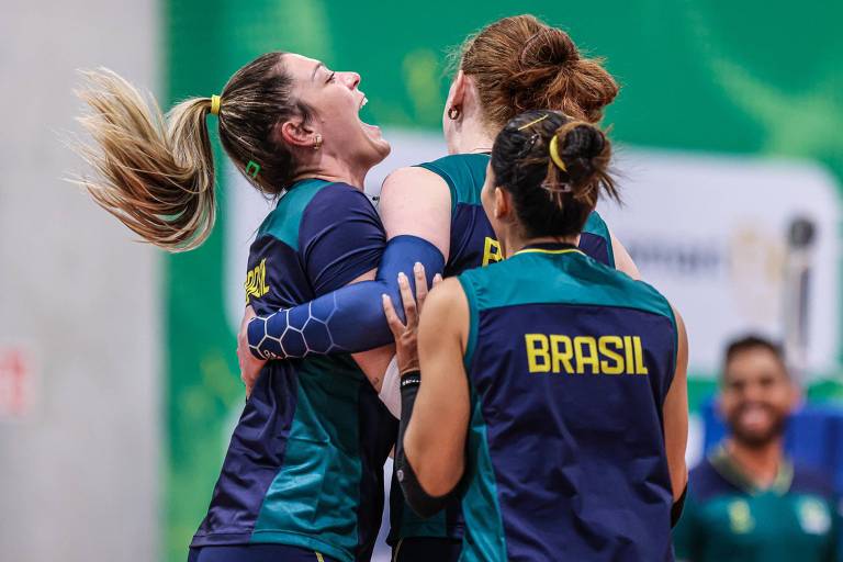 A imagem mostra um grupo de jogadoras de vôlei em um momento de celebração. Elas estão se abraçando e sorrindo, demonstrando alegria. As jogadoras vestem uniformes com as cores verde e azul, e uma delas tem o nome 'BRASIL' visível nas costas.