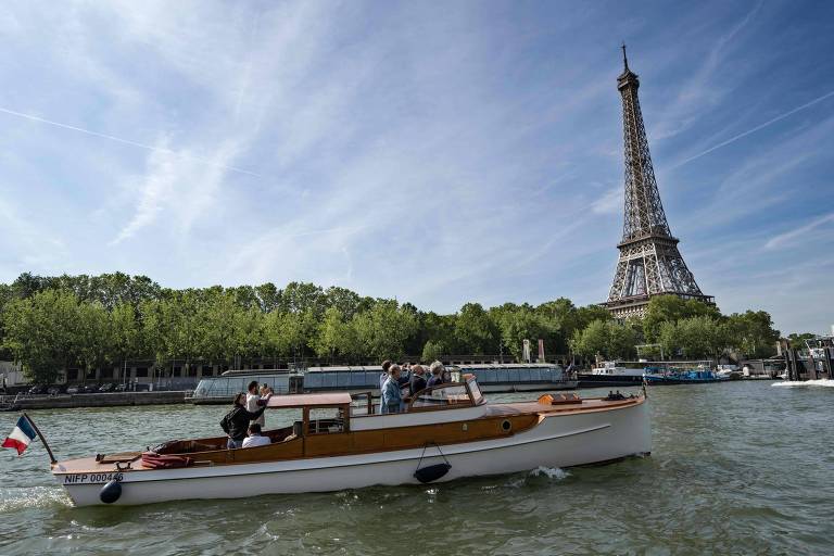 A imagem mostra um barco de madeira navegando no rio Sena, com a Torre Eiffel ao fundo. O céu está claro e azul, com algumas nuvens. O barco tem uma bandeira francesa e várias pessoas a bordo, algumas delas tirando fotos. À margem do rio, há árvores verdes e outros barcos ancorados.
