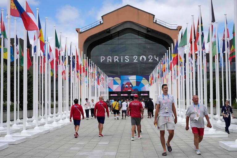A imagem mostra a entrada de um local dos Jogos Olímpicos de Paris 2024, com um grande edifício ao fundo. O edifício tem a inscrição 'PARIS 2024' em destaque. Ao longo do caminho, há várias bandeiras de diferentes países dispostas em fileiras. Algumas pessoas estão caminhando em direção à entrada, vestindo roupas casuais.
