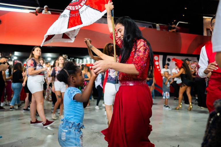 A imagem mostra um ambiente festivo onde várias pessoas estão dançando. No primeiro plano, uma mulher com uma blusa vermelha brilhante segura uma bandeira enquanto dança com uma menina pequena, que usa uma blusa azul e shorts. Ao fundo, há outras pessoas dançando e se divertindo, com um ambiente decorado em tons de vermelho e preto.