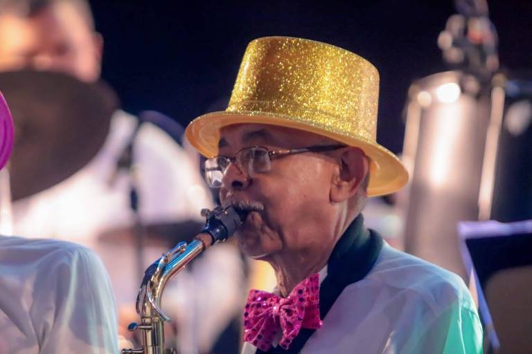 Um músico idoso está tocando saxofone. Ele usa um chapéu dourado brilhante e uma gravata borboleta rosa. Ao fundo, é possível ver outros instrumentos musicais e músicos. A iluminação é suave, criando um ambiente festivo.