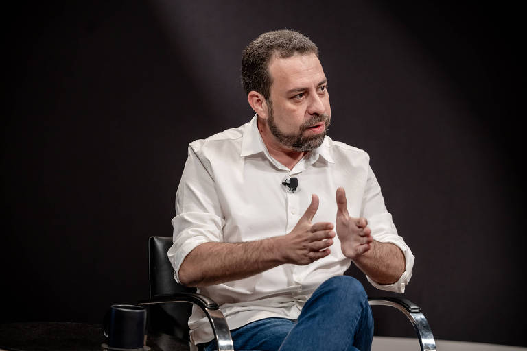 Um homem sentado em uma cadeira, usando uma camisa branca e jeans. Ele está gesticulando com as mãos enquanto fala, em um ambiente com fundo escuro. 