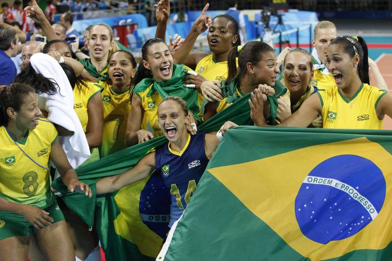 Um grupo de jogadoras da seleção brasileira de vôlei celebra uma vitória, segurando a bandeira do Brasil. Elas estão vestidas com uniformes amarelos e verdes, sorrindo e demonstrando alegria. Algumas jogadoras estão levantando as mãos, enquanto outras seguram a bandeira. O ambiente parece ser uma quadra de vôlei, com público ao fundo.