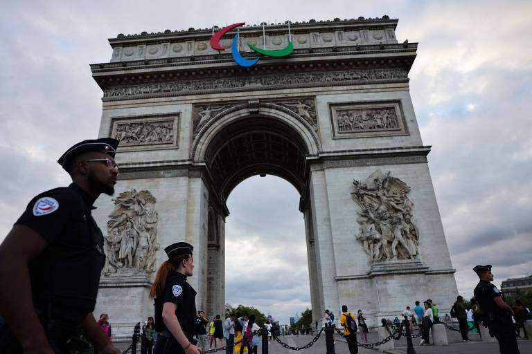 A imagem mostra o Arco do Triunfo em Paris, com a bandeira dos Jogos Paralímpicos visível no topo. Dois policiais em uniforme estão em primeiro plano, um deles com óculos escuros. Ao fundo, há várias pessoas, algumas tirando fotos, e o céu está nublado.
