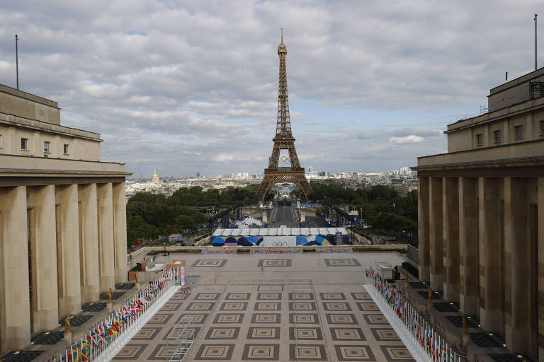 A imagem mostra uma vista da Torre Eiffel em Paris, com um céu nublado ao fundo. A torre está centralizada na imagem, e há uma grande praça em primeiro plano, cercada por edifícios. Bandeiras coloridas estão dispostas ao longo da praça