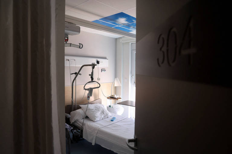 A imagem mostra a entrada de um quarto de hospital, com o número 304 visível na porta. Dentro do quarto, há uma cama hospitalar com lençóis brancos e um travesseiro. Equipamentos médicos estão visíveis ao lado da cama, incluindo um suporte para soro. A iluminação é suave e há uma janela ao fundo, permitindo a entrada de luz natural.