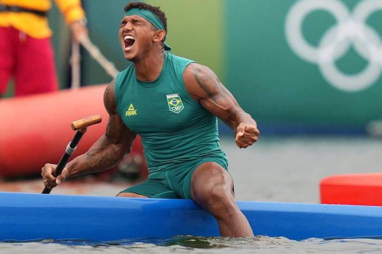 Um atleta de canoagem está em uma canoa azul, sentado e gritando de emoção. Ele usa uma camiseta verde e um boné. Ao fundo, há boias de competição e o símbolo dos Jogos Olímpicos. A água está agitada e há outros competidores em canoas vermelhas.