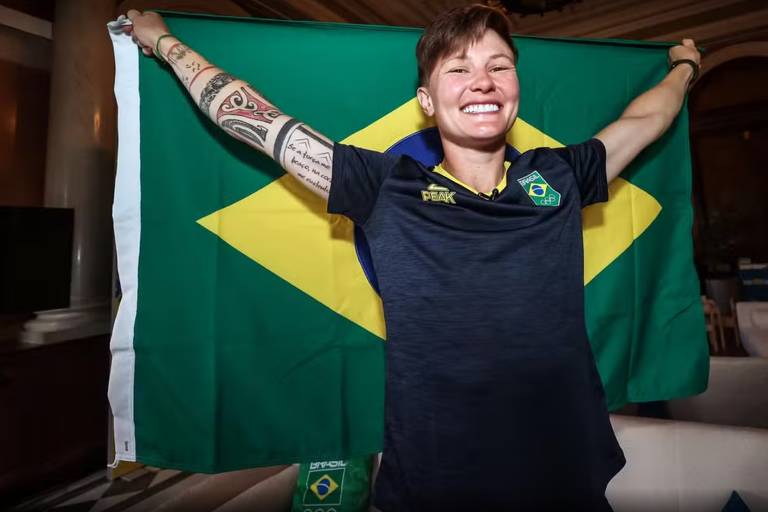 Uma atleta sorri enquanto segura a bandeira do Brasil, que possui um grande losango amarelo em um fundo verde. Ela está vestindo uma camiseta escura e tem tatuagens visíveis nos braços. O ambiente parece ser interno, com móveis ao fundo.