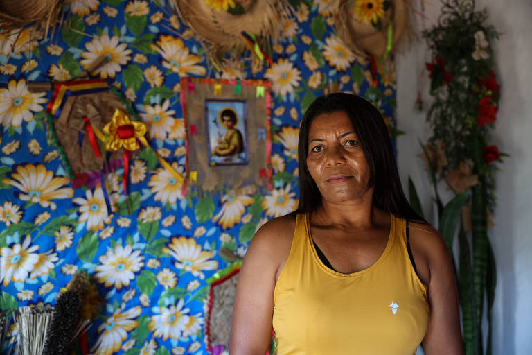 A imagem mostra uma mulher de pele morena, com cabelo longo e solto, vestindo uma blusa amarela. Ela está em um ambiente decorado com um fundo de flores brancas e azuis. Ao fundo, há uma imagem religiosa e elementos decorativos como chapéus e plantas.