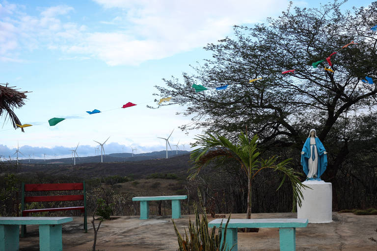 A imagem mostra uma área externa com uma estátua da Virgem Maria em pé sobre um pedestal branco. Ao fundo, há um campo com turbinas eólicas visíveis. A cena é decorada com bandeirinhas coloridas penduradas entre as árvores. Existem bancos de madeira vermelha e mesas em um espaço aberto, cercado por vegetação e um céu parcialmente nublado.