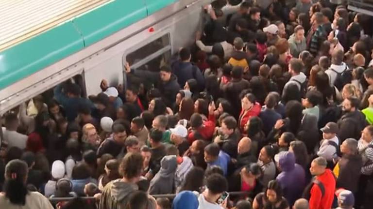 A imagem mostra uma grande multidão de pessoas tentando entrar em um trem de metrô. As pessoas estão muito próximas umas das outras, algumas segurando as portas do trem, enquanto outras parecem estar se esforçando para entrar