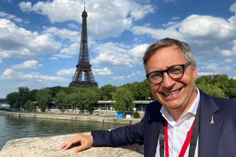 Um homem sorridente está posando para a foto com a Torre Eiffel ao fundo. Ele usa óculos e um paletó escuro, com uma camiseta branca por baixo. O céu está parcialmente nublado, e a cena inclui árvores e o rio próximo à torre.