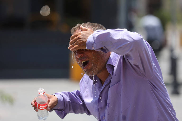 Homem de camisa lilás joga água no rosto; ele segura uma garrafa plástica vazia com rótulo da Coca-Cola