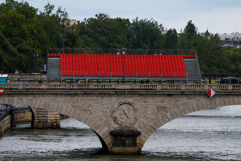A imagem mostra uma ponte de pedra sobre um rio, com uma estrutura em cima coberta por cadeiras vermelhas. Ao fundo, há árvores e um céu nublado. A ponte possui um detalhe circular no centro e bandeiras nas extremidades.
