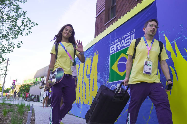 A imagem mostra dois indivíduos caminhando em direção à câmera. A mulher à esquerda, com um skate na mão, usa uma camiseta amarela e calças roxas, sorrindo e acenando. O homem à direita, também vestido com camiseta amarela e calças roxas, carrega uma mala preta e parece concentrado. Ao fundo, há um mural com a bandeira do Brasil e a inscrição 'COMITÊ OLÍMPICO DO BRASIL'.
