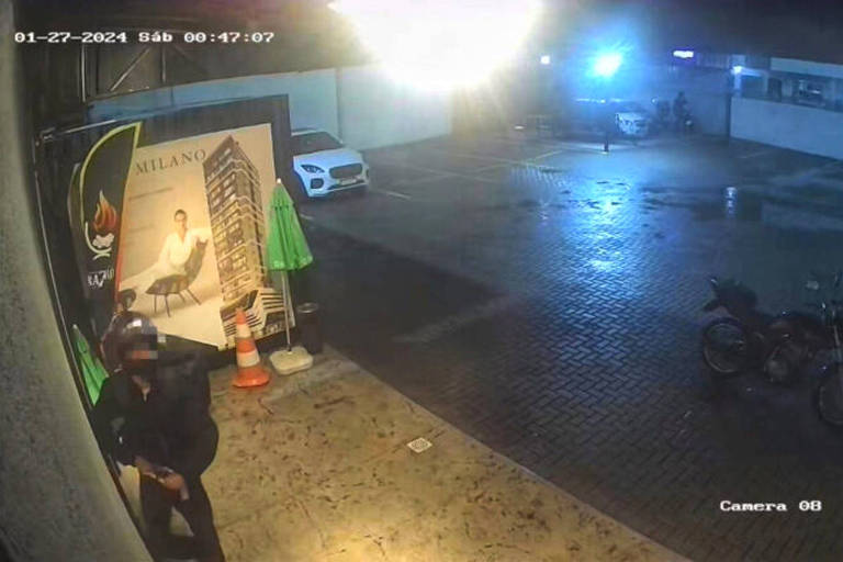 Na foto, a imagem de uma câmera de segurança registra uma área externa vizinha a um estacionamento, e uma pessoa vestida de entregador entrando em um estabelecimento. Ao lado, uma motocicleta estacionada.