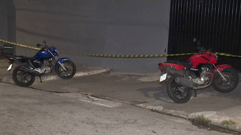 A imagem mostra duas motocicletas estacionadas em uma calçada. A motocicleta à esquerda é de cor azul e a da direita é vermelha. Ambas estão próximas a uma parede cinza e estão cercadas por fita de isolamento amarelo e preto