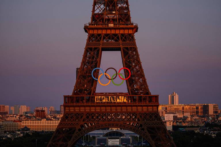 A imagem mostra a Torre Eiffel em Paris ao entardecer, com os cinco anéis olímpicos coloridos (azul, amarelo, preto, verde e vermelho) pendurados na estrutura. O céu apresenta um tom suave de roxo, e ao fundo, é possível ver a cidade de Paris com edifícios iluminados.
