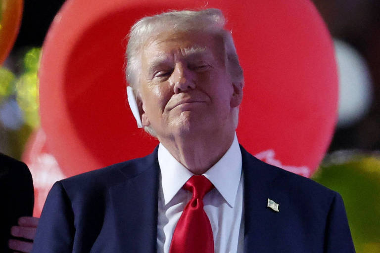 A imagem mostra Donald Trump, homem com cabelo loiro, usando um terno escuro e uma gravata vermelha. Ele está sorrindo levemente e parece estar em um evento, com balões coloridos ao fundo, incluindo um grande balão vermelho.
