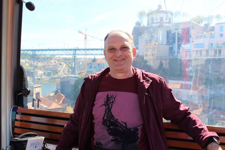 Um homem sorridente está sentado em um teleférico, com uma vista panorâmica da cidade do Porto ao fundo. O teleférico é de vidro, permitindo uma visão clara da paisagem, que inclui um rio e uma ponte