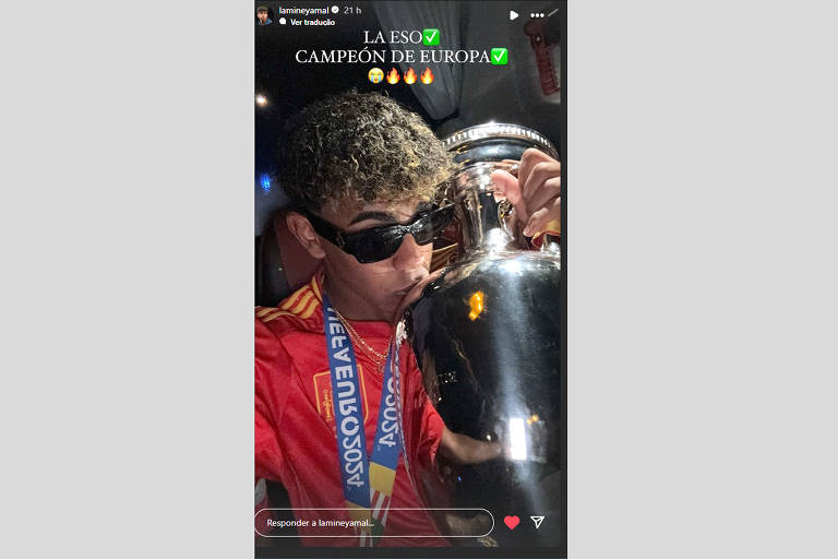 Selfie mostra Lamine Yamal beijando a taça da Eurocopa. Lamine é um garoto negro com cabelos cacheados com pontas loiras. Ele usa um óculos escuro, a camisa vermelha da sua seleção e a medalha de campeão. Na foto tem o escrito "La ESO" com um símbolo verde na frente e "Campeão da Europa" com o mesmo emoticon na frente.