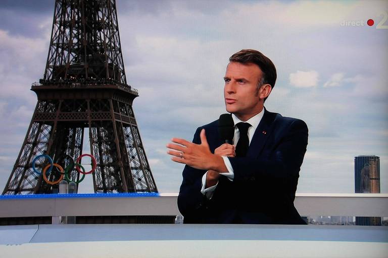 A imagem mostra o presidente francês Emmanuel Macron em um terno escuro sentado em uma mesa, gesticulando com as mãos. Ao fundo, é visível a Torre Eiffel sob um céu nublado. O homem parece estar em uma entrevista ou apresentação.