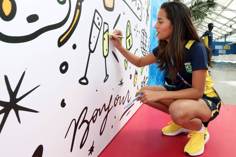 Agachada, Rayssa Leal, medalhista de prata no skate street em Tóquio-2020, usa a mão direita para colorir com tinta amarela um painel em Saint-Ouen, na França, antes dos Jogos Olímpicos