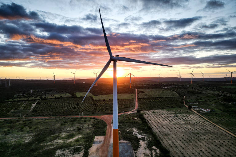 A imagem mostra um parque eólico com várias turbinas eólicas, sob um céu colorido durante o pôr do sol. Há uma estrada de terra ao fundo da paisagem.