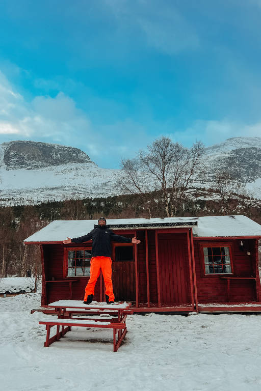 Um homem está em pé sobre uma mesa vermelha na frente de uma cabana vermelha, cercada por neve. Ele usa calças laranjas e uma jaqueta escura, com os braços abertos e olhando para o céu. Ao fundo, há montanhas cobertas de neve sob um céu azul com algumas nuvens.