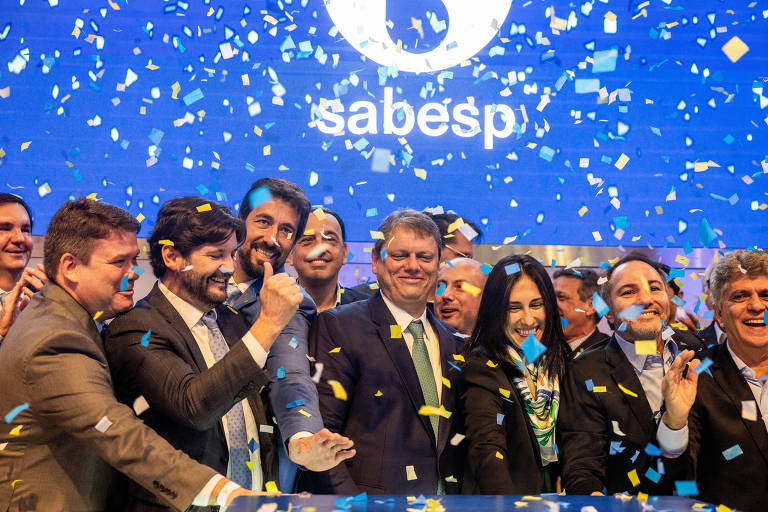 Um grupo de pessoas aparece sorridente em meio a uma chuva de papéis picados. No centro está o governador de São Paulo, Tarcísio de Freitas. Atrás das pessoas está um telão azul marinho com o símbolo da Sabesp em branco.