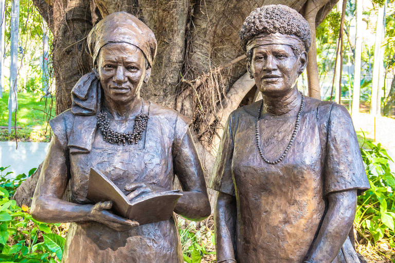 A imagem mostra duas estátuas de bronze representando mulheres. A primeira mulher está vestida com um vestido longo e segura um livro, enquanto a segunda mulher usa um vestido mais curto e segura uma pasta. Ambas estão posicionadas em frente a uma árvore grande, com vegetação ao redor. No chão, há placas informativas sobre as estátuas