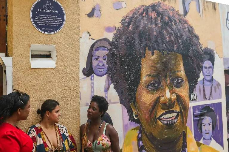 A imagem mostra um mural colorido de Lélia Gonzalez, uma importante figura na luta pelos direitos raciais e feministas no Brasil. Ao lado do mural, três mulheres estão conversando. O mural retrata Lélia com cabelo crespo e um sorriso, enquanto ao fundo há outras figuras femininas representadas