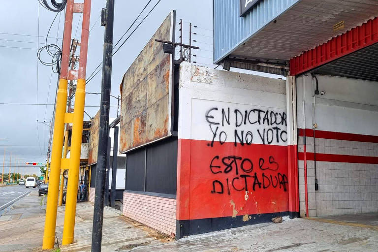 A imagem mostra um muro de um edifício com grafite em letras grandes. O texto diz: 'EN DICTADOR YO NO VOTO' e 'ESTO ES DICTADURA'. O muro é pintado em vermelho e branco, e ao fundo há uma estrutura metálica e fios elétricos visíveis. O céu está nublado.
