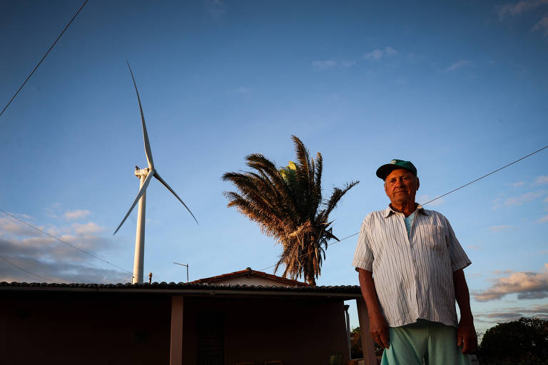 Um homem está posicionado em primeiro plano, em frente a uma casa simples. Ao fundo, há uma turbina eólica e uma palmeira. O céu apresenta um tom azul com nuvens, indicando o final do dia.
