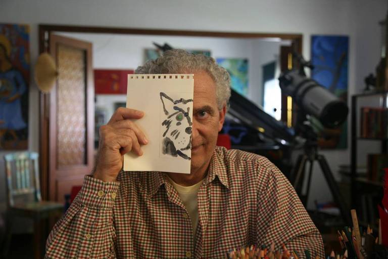 Um homem com cabelo grisalho e encaracolado está segurando uma folha de papel que mostra um desenho de um animal, possivelmente um lobo ou um cachorro, cobrindo parcialmente seu rosto. Ele usa uma camisa xadrez e está sentado em uma mesa com lápis de cor ao seu redor. Ao fundo, há uma sala decorada com várias obras de arte e um telescópio.