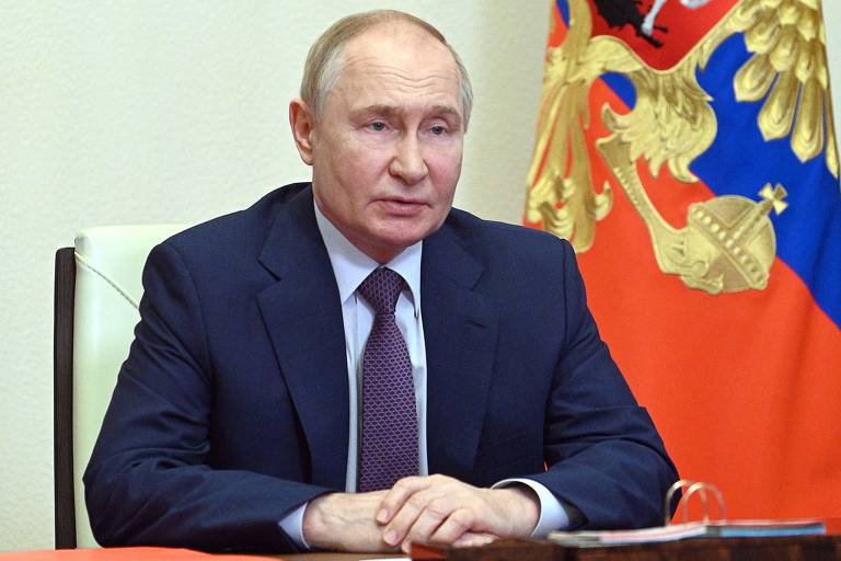 Um homem está sentado em uma mesa, vestindo um terno escuro e uma gravata. Ele parece estar em uma reunião oficial, com as mãos apoiadas na mesa. Ao fundo, há uma bandeira russa com detalhes em vermelho, azul e dourado, incluindo um brasão. A parede é de cor clara.
