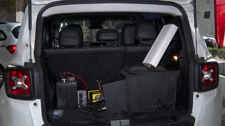 A imagem mostra o interior do porta-malas de um veículo branco. No fundo, há bancos de couro escuro. No chão, estão visíveis uma bateria, um equipamento eletrônico em uma caixa preta, e um objeto cilíndrico prateado. Também há fios conectando os dispositivos