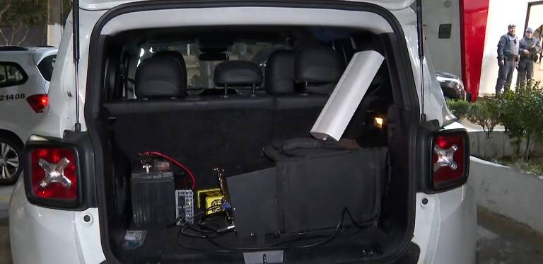 A imagem mostra o interior do porta-malas de um veículo branco. No fundo, há bancos de couro escuro. No chão, estão visíveis uma bateria, um equipamento eletrônico em uma caixa preta, e um objeto cilíndrico prateado. Também há fios conectando os dispositivos