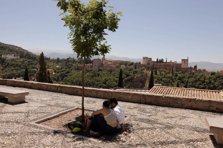 Duas pessoas sentadas no chão na sombra de uma pequena árvore; ao fundo é possível ver a Alhambra, ponto turístico da cidade