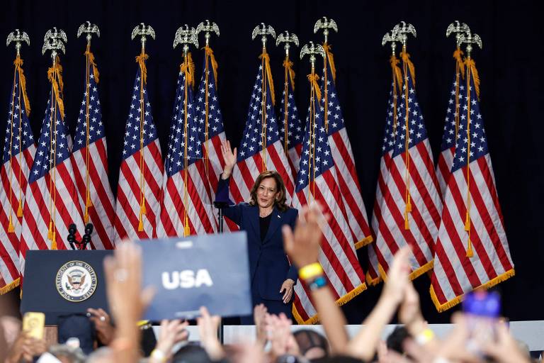 A imagem mostra uma mulher acenando para a multidão em um evento político. Ao fundo, há várias bandeiras dos Estados Unidos dispostas em fileira. A mulher está vestida com um terno escuro e está em frente a um púlpito com o selo presidencial. A multidão à sua frente levanta as mãos, algumas segurando cartazes, incluindo um que diz 'USA'.
