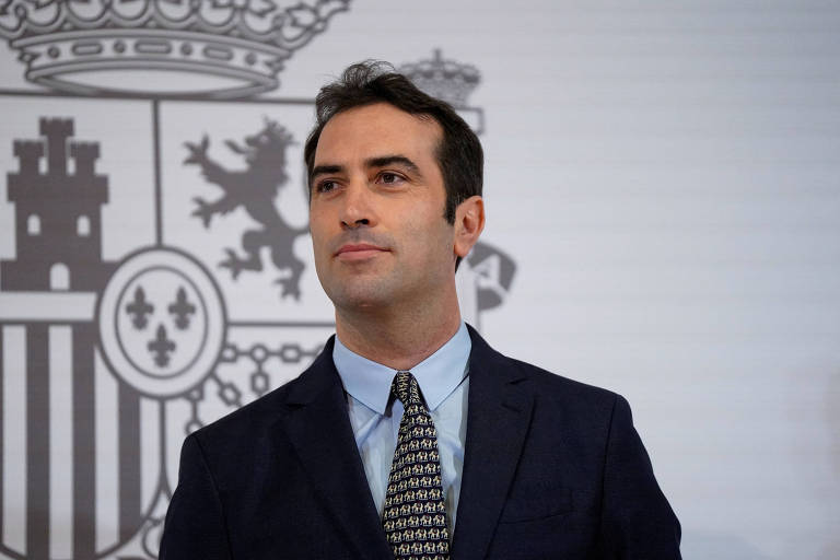 Um homem em um terno escuro e gravata com padrão, posando em frente a um fundo que apresenta símbolos da Espanha, incluindo uma coroa e brasões. Ele tem cabelo escuro e está olhando para o lado, com uma expressão séria.