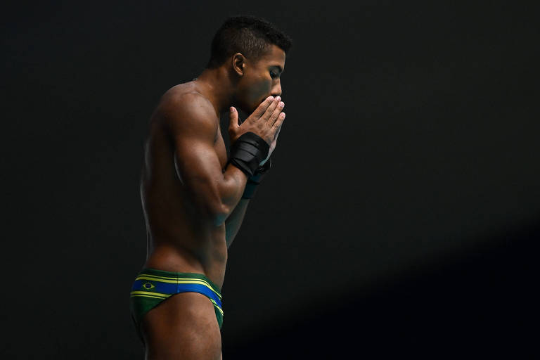 Um atleta de mergulho está em posição de preparação, com as mãos unidas em frente ao rosto. Ele é de pele morena e está usando um traje de banho com as cores da bandeira do Brasil, que inclui verde, amarelo e azul. O fundo da imagem é escuro, destacando a figura do atleta.
