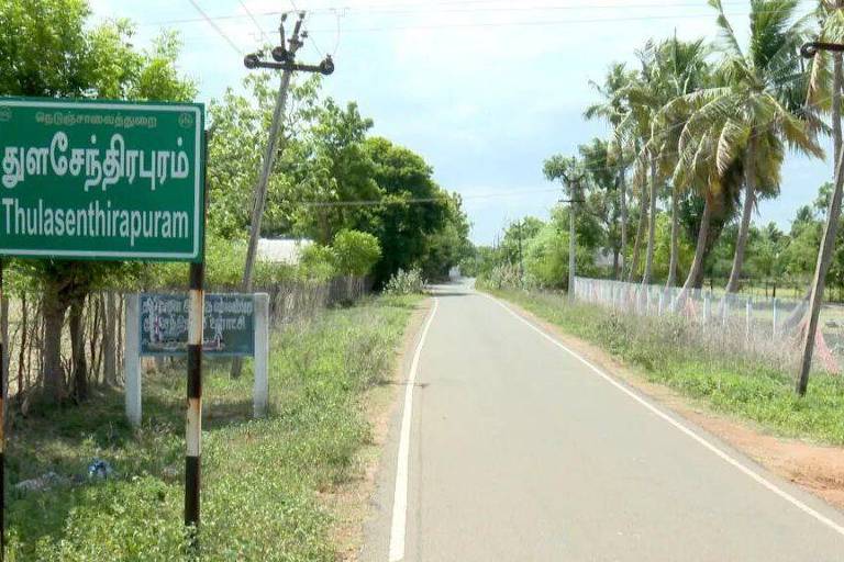 A imagem mostra uma estrada rural em Thulasenthirapuram, com uma placa verde em Tamil indicando o nome da localidade. O cenário é cercado por vegetação, incluindo árvores e arbustos, e há uma cerca ao fundo. O céu está parcialmente nublado.