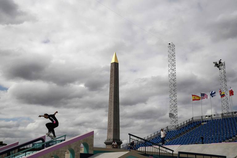 A imagem mostra um skatista realizando uma manobra em uma rampa, com um obelisco ao fundo. O obelisco tem uma ponta dourada e está cercado por nuvens escuras. À direita, há torres de iluminação e várias bandeiras de diferentes países. A arquibancada é composta por assentos azuis.
