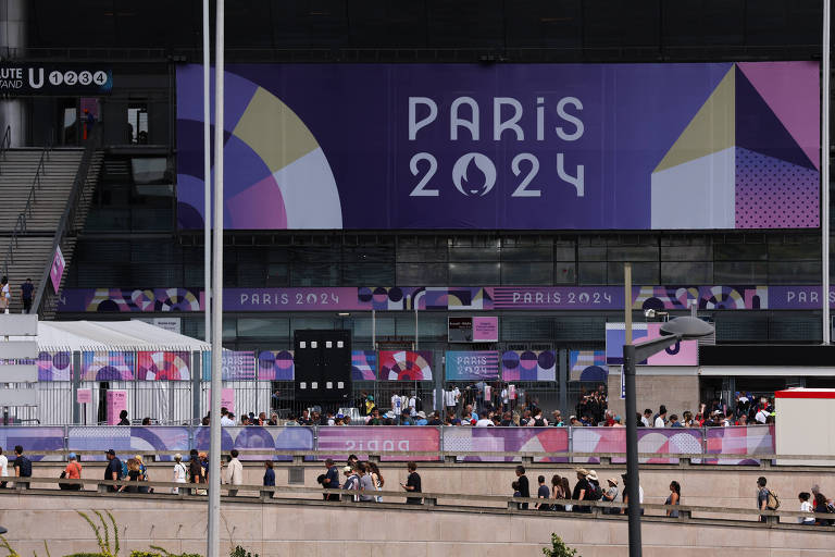 A imagem mostra um grande painel com a inscrição 'PARIS 2024', decorado com formas geométricas em cores variadas, como roxo, azul e amarelo. Abaixo do painel, há uma multidão de pessoas caminhando em direção ao evento, com barracas e estruturas montadas ao fundo.
