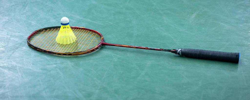 A imagem mostra uma raquete de badminton posicionada sobre uma superfície verde, com uma peteca amarela em cima da rede da raquete. A raquete tem um cabo preto e uma estrutura metálica com cordas visíveis.